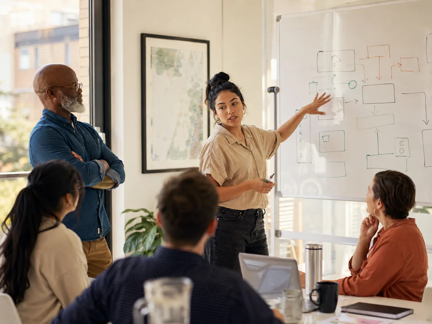 A Latina team lead at a whiteboard mid-gesture, confidently walking a diverse team through a framework in a bright room