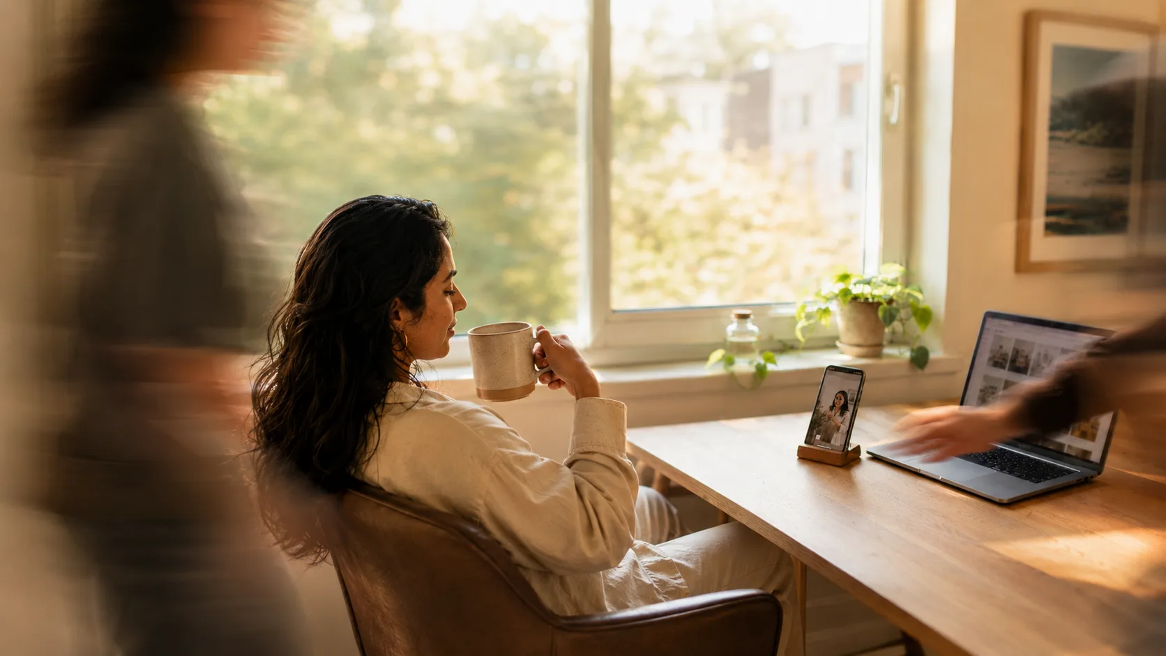A woman leaning back in her chair with a coffee, calm and in sharp focus, watching one of her finished clips play on a phone propped beside a laptop full of them, while the room blurs into motion around her